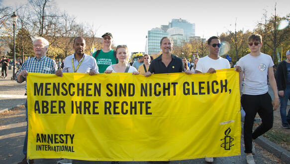 Eine Gruppe von mehreren Personen geht auf einer Straße und hält ein Banner hoch, auf dem steht: "Menschen sind nicht gleich. Aber ihre Rechte".